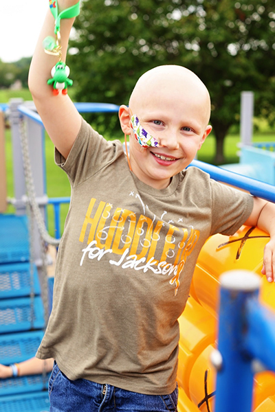 Smiling young boy with a medical tube taped to his cheek, wearing a custom fundraiser t-shirt that reads “Huddle up  for Jackson,” standing on playground. Image represents CAUSETEAM’s medical t-shirt fundraising suppor